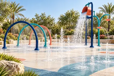 AI concept visual: a dedicated splash pad in a bright landscaped park, fine ground water jets, three low curved spray arches and a small tipping bucket feature, navy and teal posts, restrained.